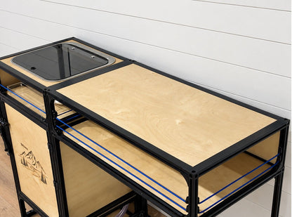 A modular campervan sink and countertop galley shelf with light natural wood panels, and a black sink basin on top, photographed in a real interior setting with a white paneled wall and wood floor.