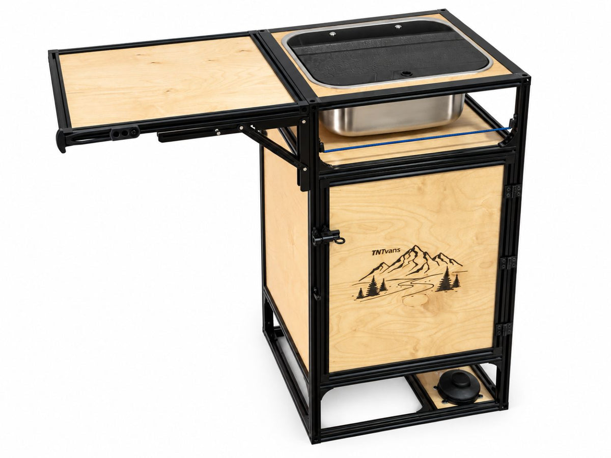 A mobile kitchen unit with a black metal frame and light natural wood panels, featuring an attached fold-down table, a sink basin with a metal pot, and a mountain graphic printed on the front door.